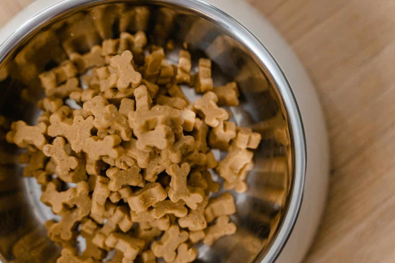 Metal bowl filled with small, bone-shaped dog treats, placed on a wooden surface, treats are light brown and uniform in size, likely prepared for feeding a pet