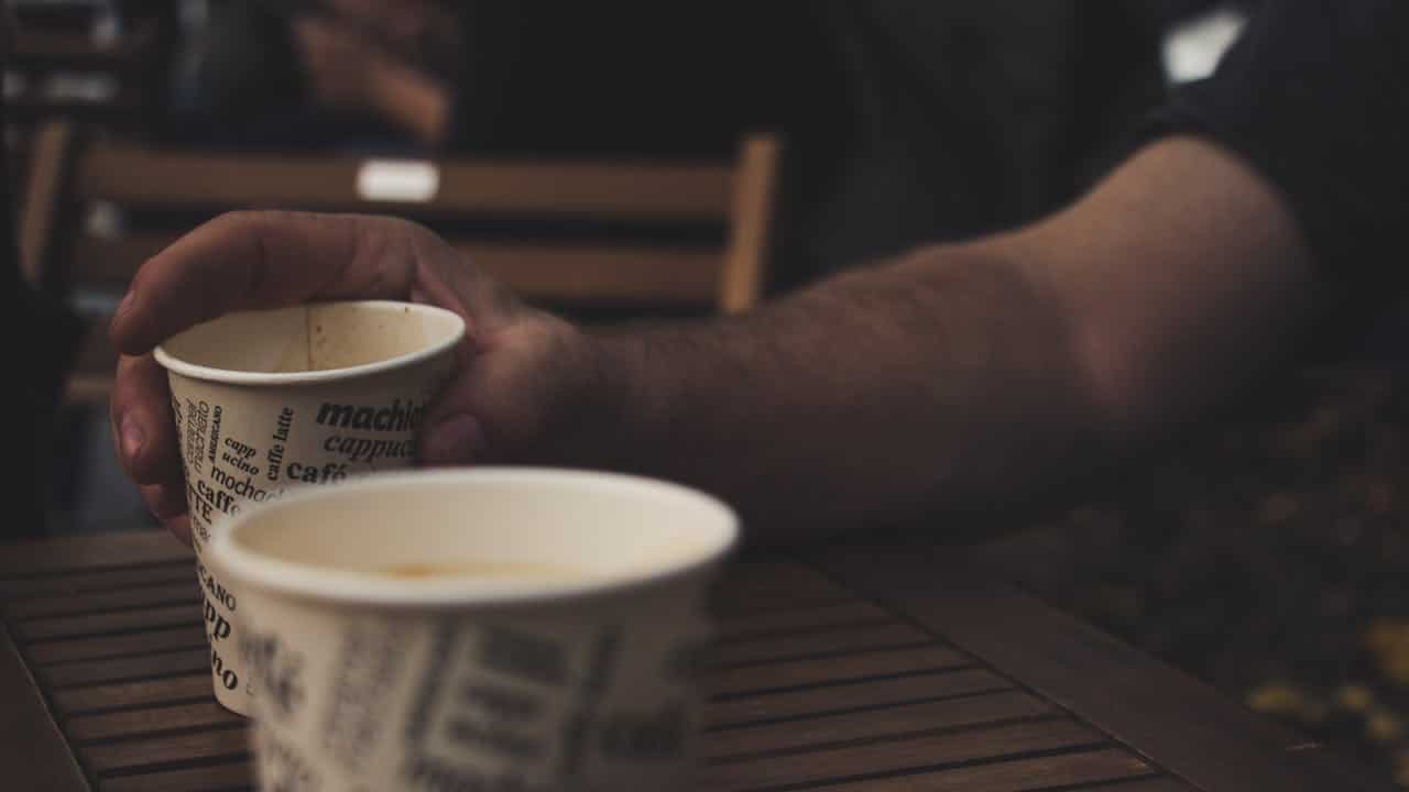 A person holding a coffee cup with the word "coffee" printed on it, with another cup visible in the foreground, sitting on a wooden table