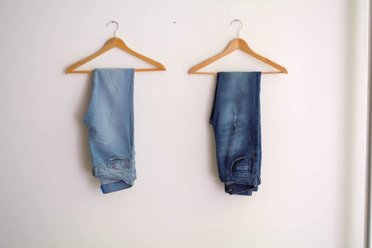 Two pairs of jeans are hanging on wooden hangers against a white wall, one pair is light blue and the other is dark blue, both pairs of jeans are neatly hung and organized, the image depicts a simple clothing display in a clean, minimal setting