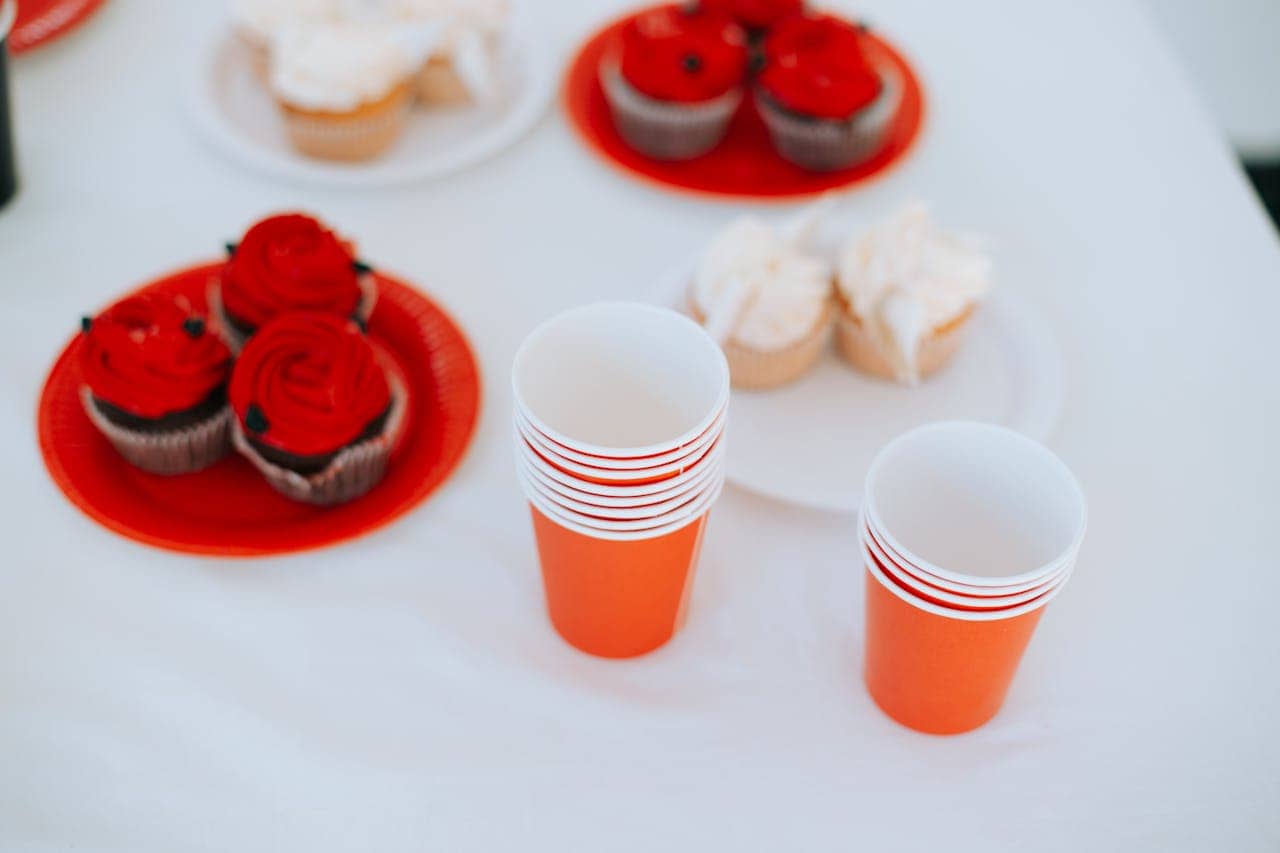 Orange disposable cups stacked neatly beside plates of red-iced cupcakes, a light, festive scene with snacks prepared for a gathering