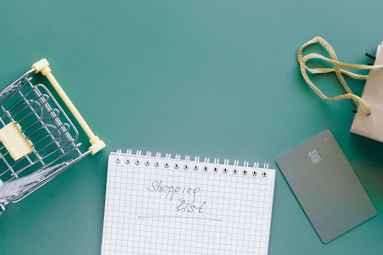 Shopping cart, shopping list, notepad with "Shopping List" written, credit card, paper shopping bag, flat lay, items for shopping, preparing to shop, organized flat composition, grocery planning, shopping essentials