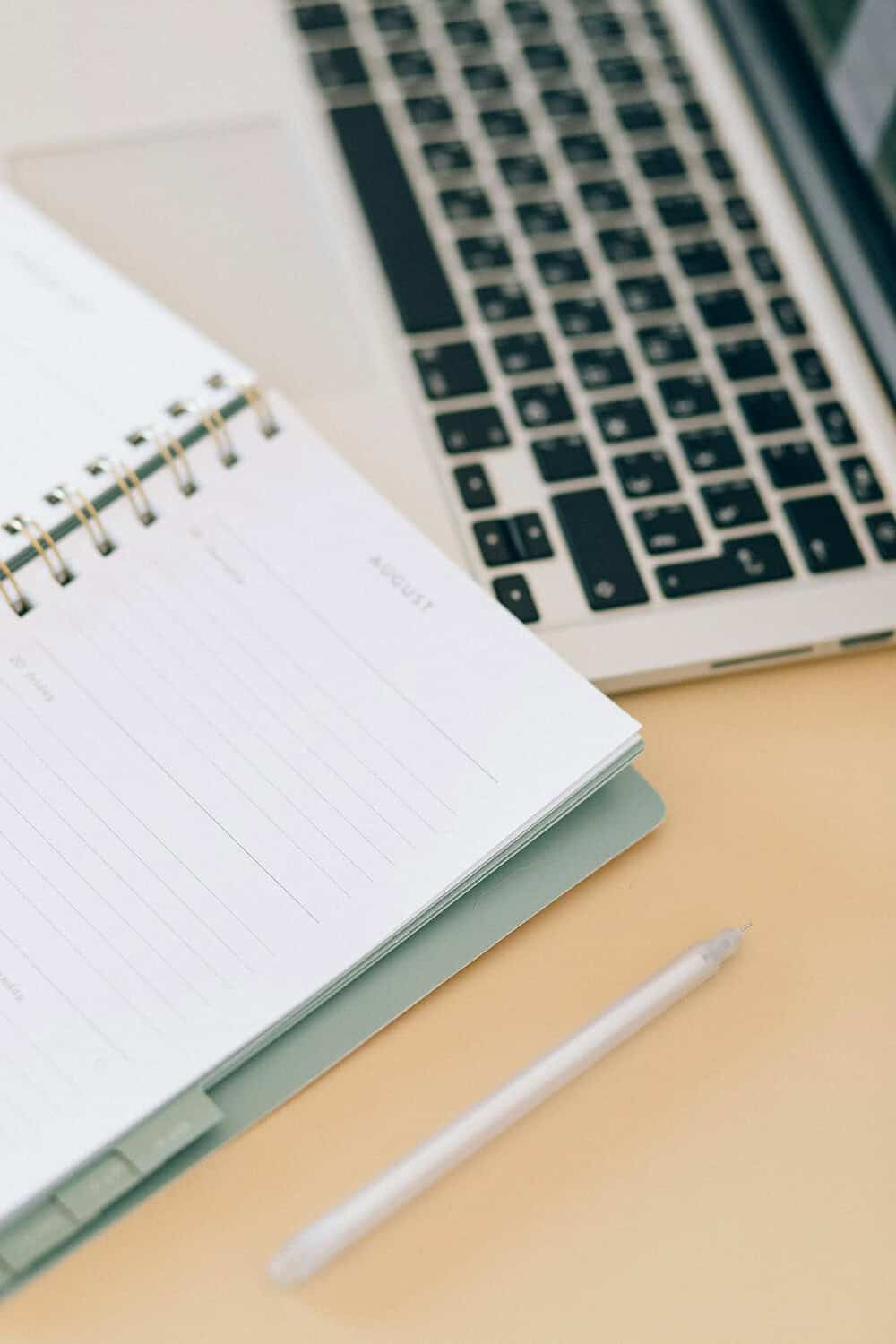 A spiral-bound planner open to the month of August, a silver laptop keyboard partially visible, a transparent pen resting on a beige desk, organized workspace setup, ideal for planning cleaning tasks
