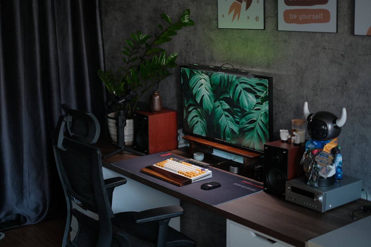 Home office desk with computer monitor displaying tropical leaves, custom keyboard, office chair, plants, and collectible figurine