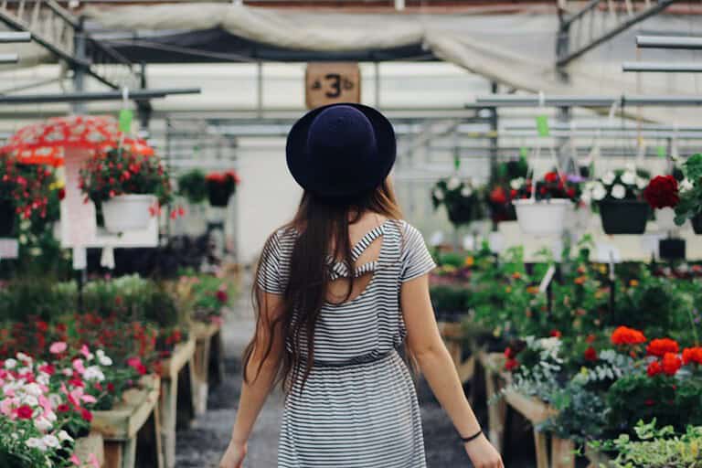 Woman in striped dress and black hat, walking in a flower shop, surrounded by colorful flowers in pots, plants hanging from above, bright indoor lighting