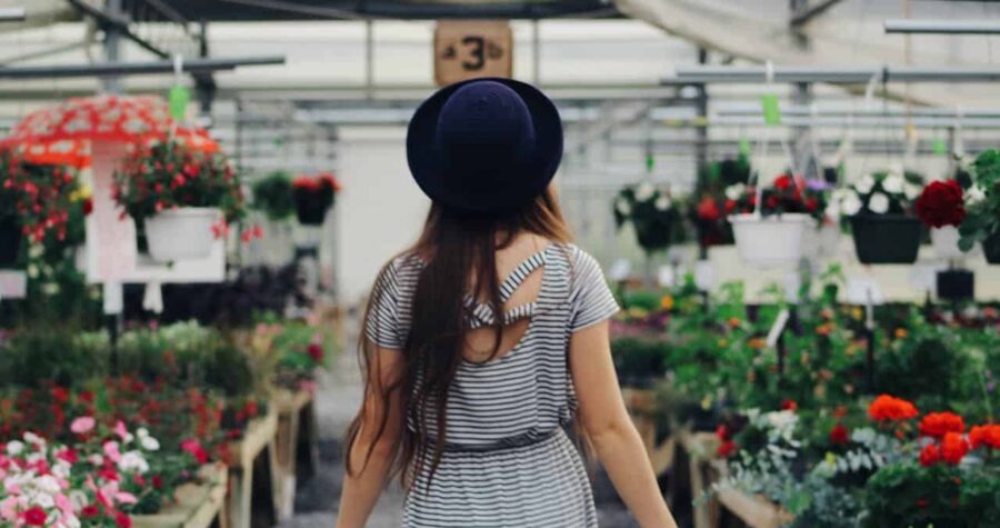 Woman in striped dress and black hat, walking in a flower shop, surrounded by colorful flowers in pots, plants hanging from above, bright indoor lighting