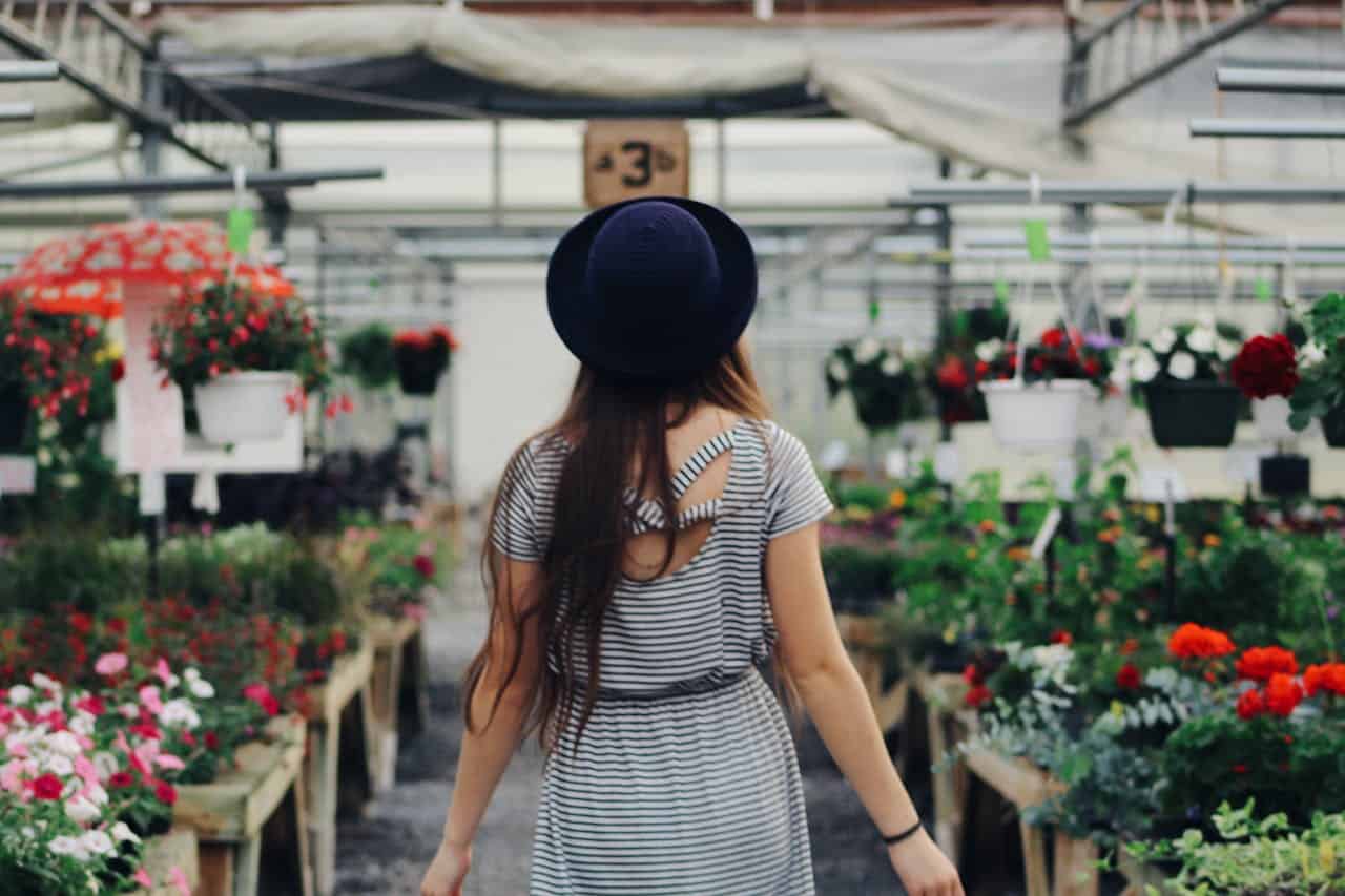 Woman in striped dress and black hat, walking in a flower shop, surrounded by colorful flowers in pots, plants hanging from above, bright indoor lighting