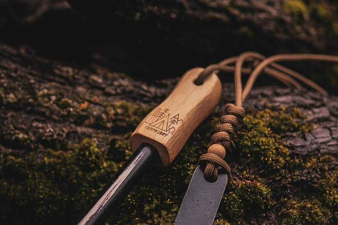 Wooden-handled firestarter tool with metal striker on moss-covered surface, featuring camping logo and paracord lanyard attachment