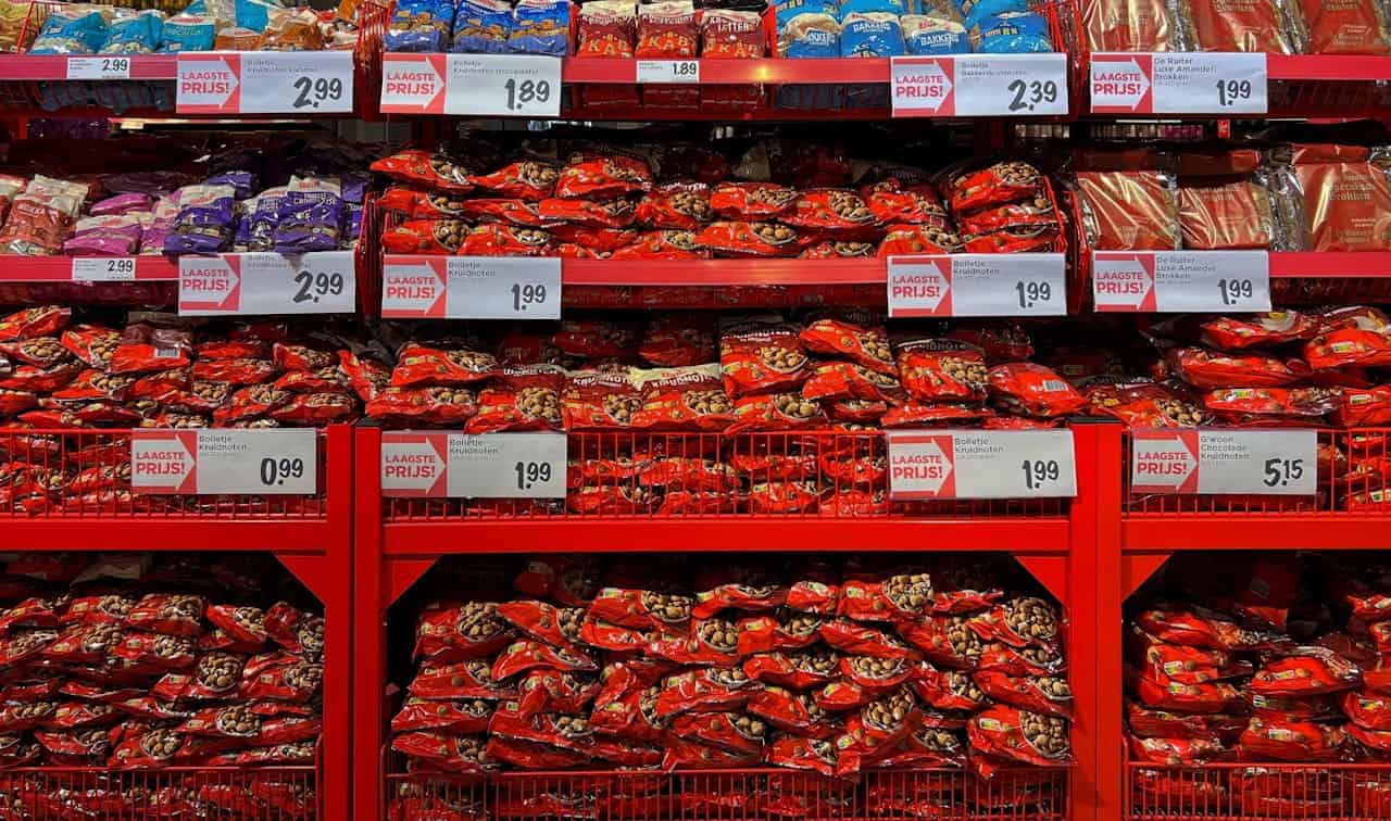 A store shelf filled with bags of snacks, red packaging with various product types, prices displayed in Dutch on signs, a variety of flavored snacks, multiple rows of items for sale, promotional signage with "LAAGSTE PRIJS" (Lowest Price)