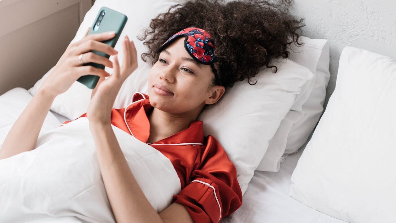 A person lying in bed, wearing a red pajama set, holding a smartphone, wearing a sleep mask with a floral pattern, smiling while looking at the phone, surrounded by white pillows and a cozy bedroom atmosphere