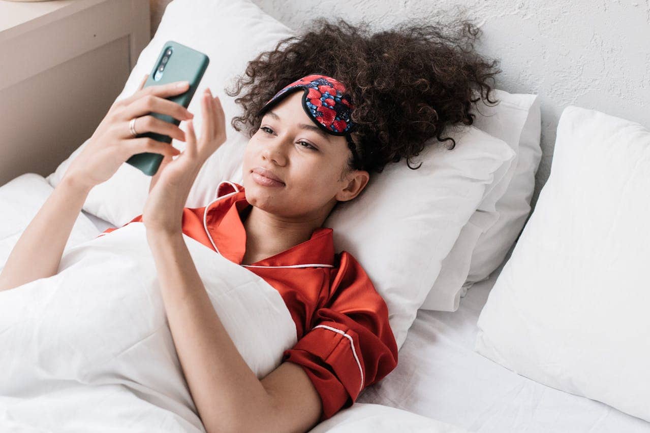 A person lying in bed, wearing a red pajama set, holding a smartphone, wearing a sleep mask with a floral pattern, smiling while looking at the phone, surrounded by white pillows and a cozy bedroom atmosphere