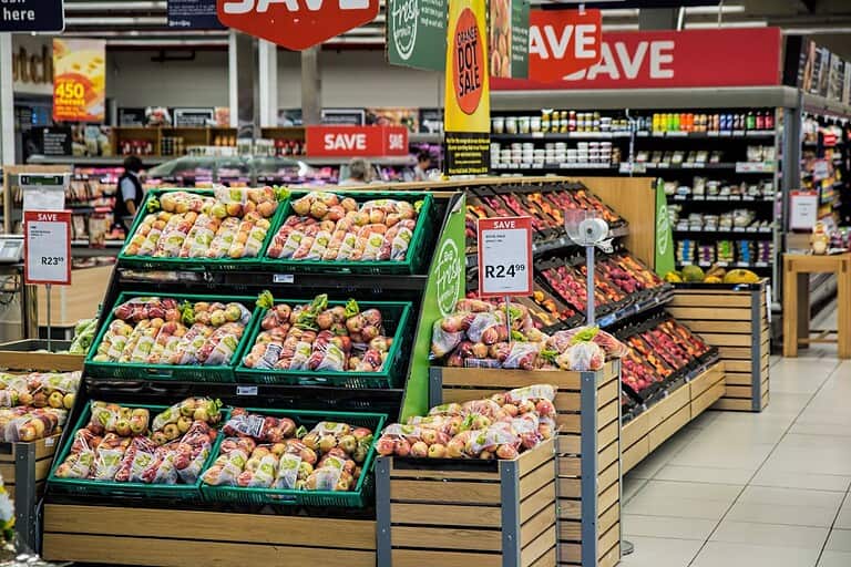 A supermarket produce section, multiple displays of apples in plastic bags, price tags showing R23.39 and R24.99, promotional signs with "SAVE," organized wooden shelves, neatly arranged fruits, vibrant fresh produce