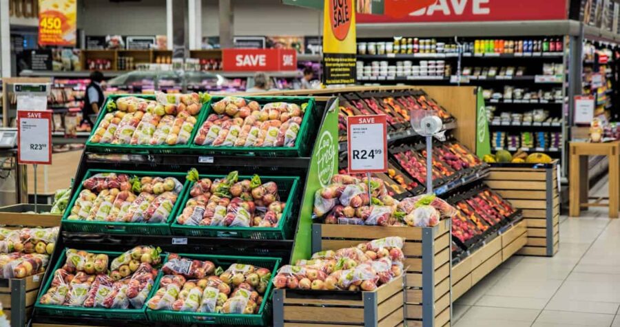 A supermarket produce section, multiple displays of apples in plastic bags, price tags showing R23.39 and R24.99, promotional signs with "SAVE," organized wooden shelves, neatly arranged fruits, vibrant fresh produce