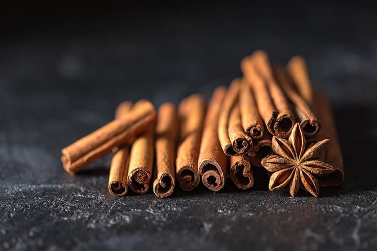 A pile of cinnamon sticks and a star anise sit on a dark surface, cinnamon sticks are arranged in a stacked pattern, star anise is placed at the center, showcasing their texture and shape
