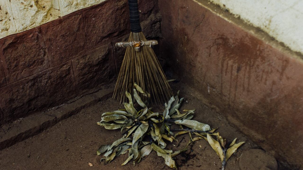 A broom resting against a wall, with a pile of dry, yellowed leaves on the ground next to it, dirt floor, textured wall with a reddish-brown base