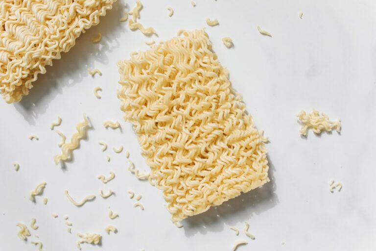 The image shows two blocks of uncooked instant ramen noodles, with some scattered pieces around them, showcasing the texture of the noodles on a white surface, emphasizing their wavy pattern
