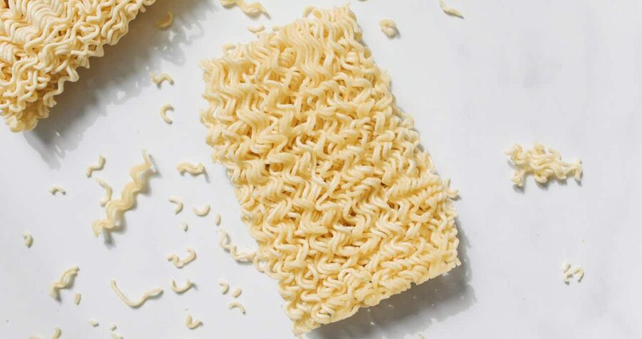The image shows two blocks of uncooked instant ramen noodles, with some scattered pieces around them, showcasing the texture of the noodles on a white surface, emphasizing their wavy pattern