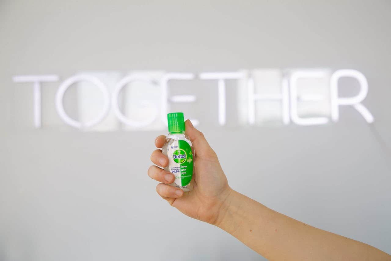 Hand holding a small bottle of Dettol hand sanitizer, clear liquid inside the bottle, green cap visible, background features the word "TOGETHER" in neon lights, focus on hygiene and cleanliness