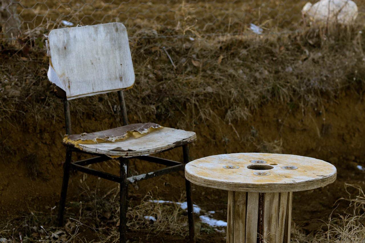 An old, worn-out chair with peeling paint, a weathered wooden spool table nearby, both placed outdoors, dirt and dry grass surrounding them, visible natural wear and tear