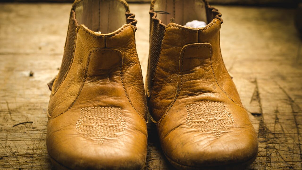 A pair of well-worn brown leather shoes, the shoes have a worn texture and detailed stitching, displayed on a wooden surface, showing a rustic, aged appearance
