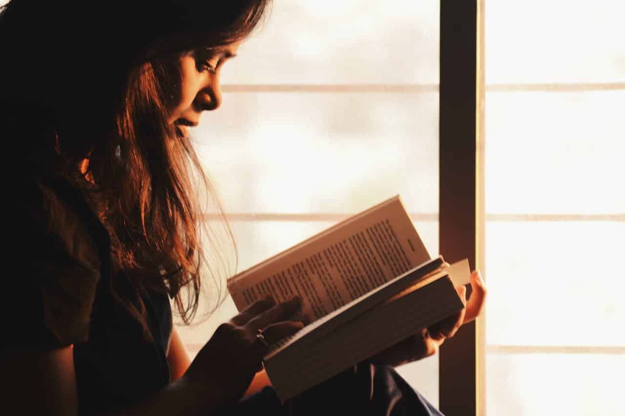Woman reading a book, sitting by a window, soft natural light illuminating her face, focused and absorbed in reading