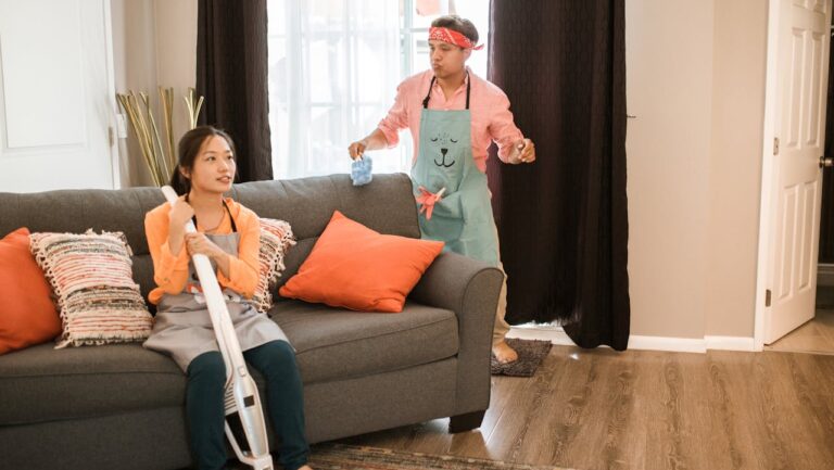 A woman sitting on a couch holding a vacuum cleaner, wearing an apron, man standing nearby holding a cleaning cloth, both dressed casually, playful mood, cleaning scene