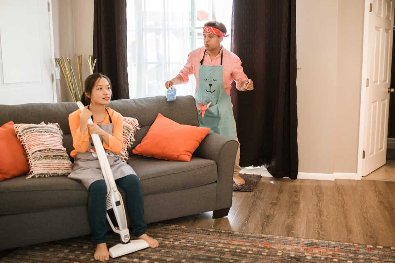 A woman sitting on a couch holding a vacuum cleaner, wearing an apron, man standing nearby holding a cleaning cloth, both dressed casually, playful mood, cleaning scene