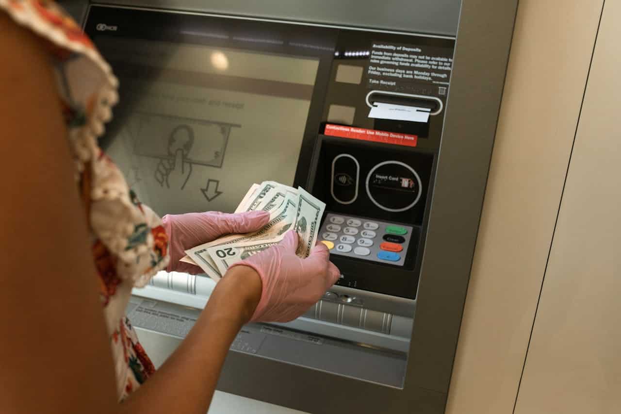 A person wearing pink gloves, holding several $20 bills, preparing to deposit cash into an ATM, with the ATM screen showing instructions for depositing money