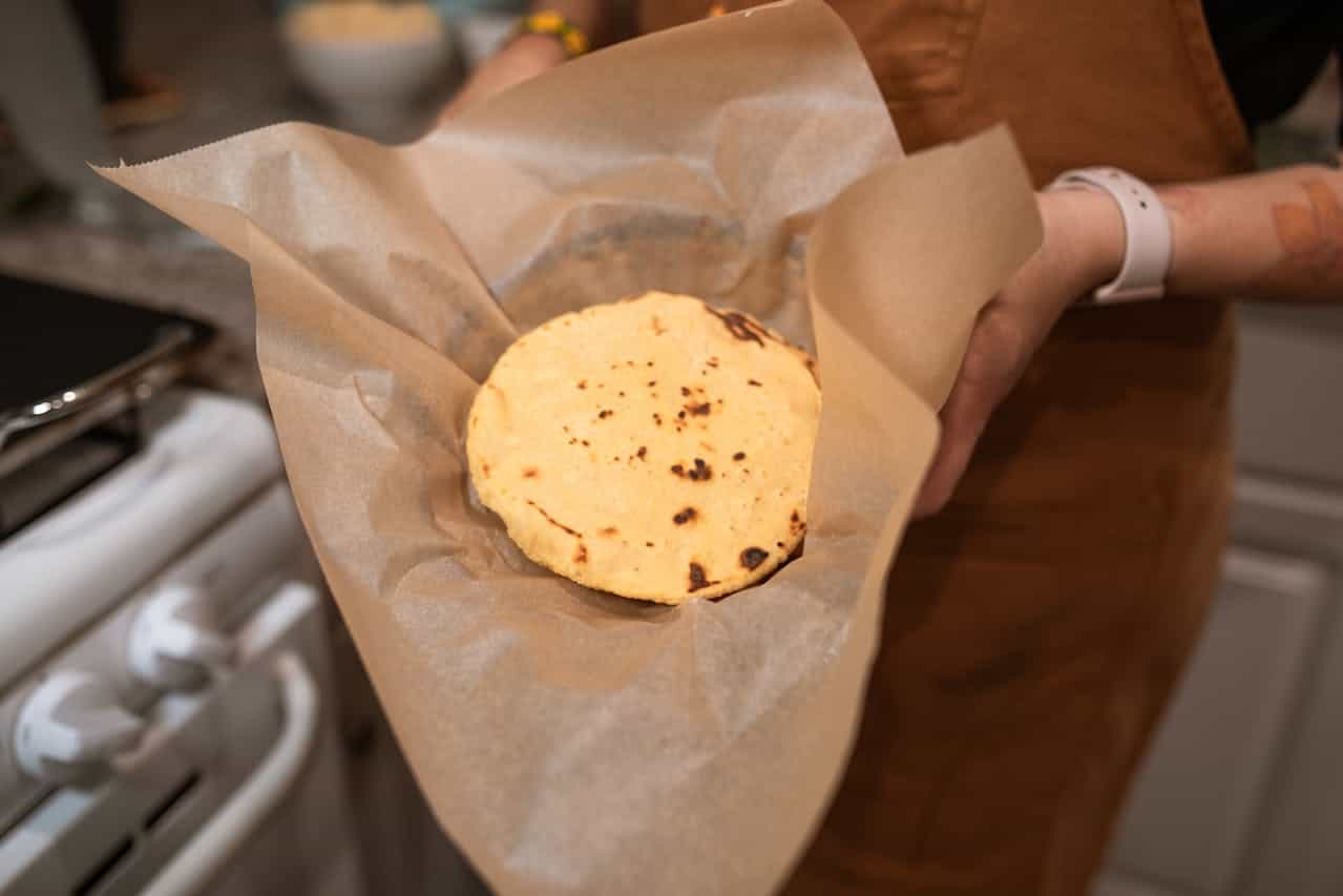 A person holding a freshly made flatbread wrapped in parchment paper, the flatbread has slightly charred spots, kitchen environment with stove visible in the background
