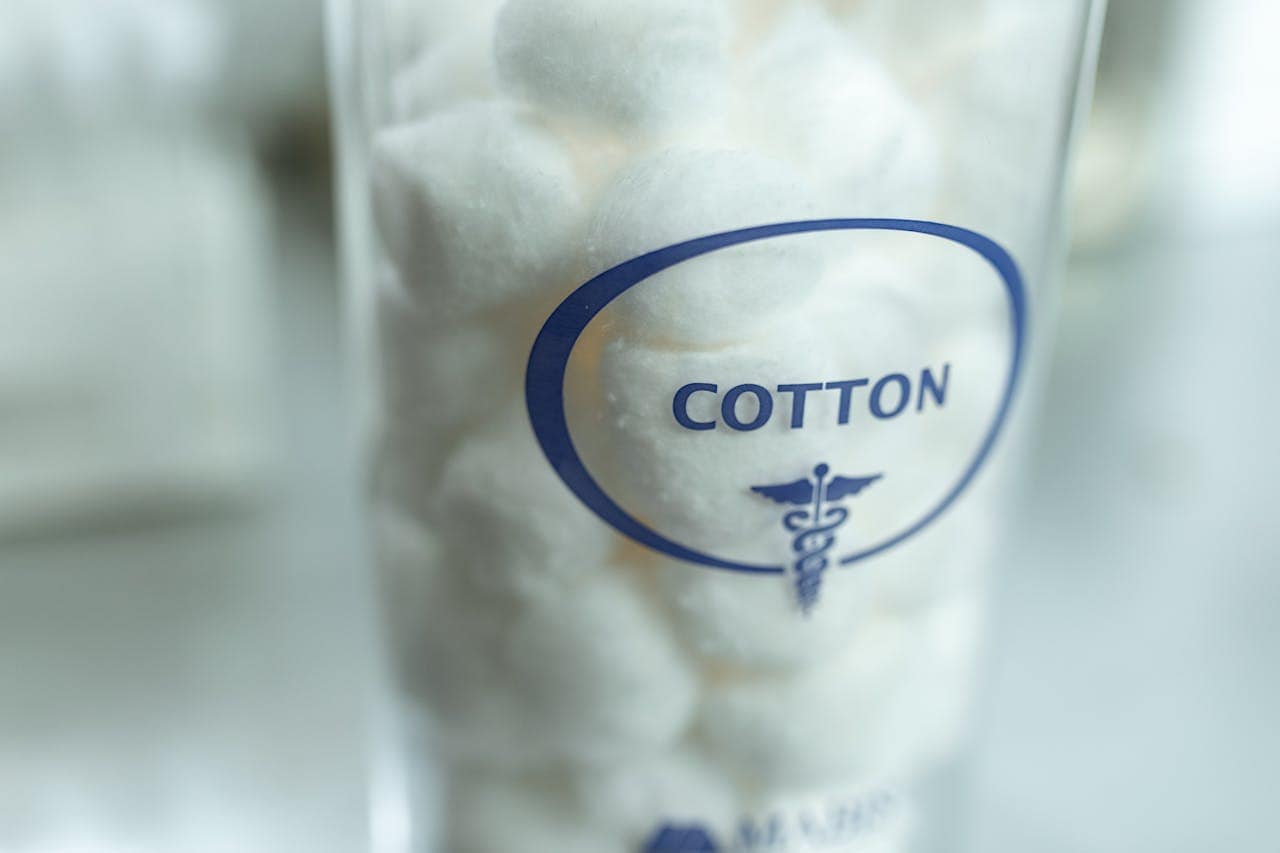 Cotton balls in a glass container, labeled "COTTON," with a medical symbol, close-up view, clear glass container with cotton inside, well-lit image, clean background, focus on cotton balls