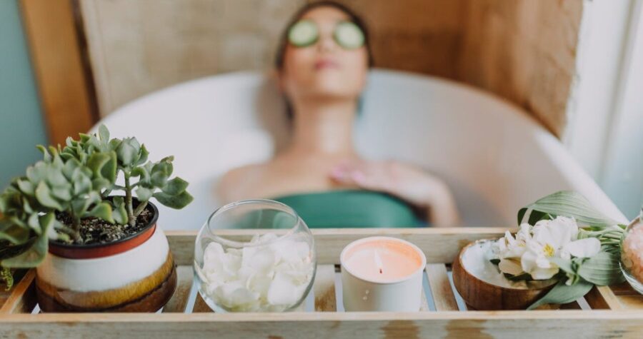 A woman relaxing in a bath with cucumber slices on her eyes, a wooden tray with a candle, rose petals, a plant, and flower petals in a bowl placed beside the bath, peaceful and serene spa-like setting, the focus on self-care and relaxation