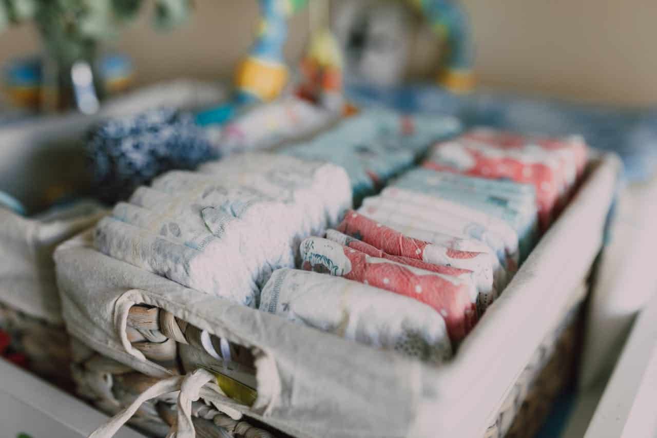 Woven basket filled with neatly stacked baby diapers, soft fabric lining tied with a bow, diapers arranged in pastel blue, white, and red patterns, organized nursery essentials