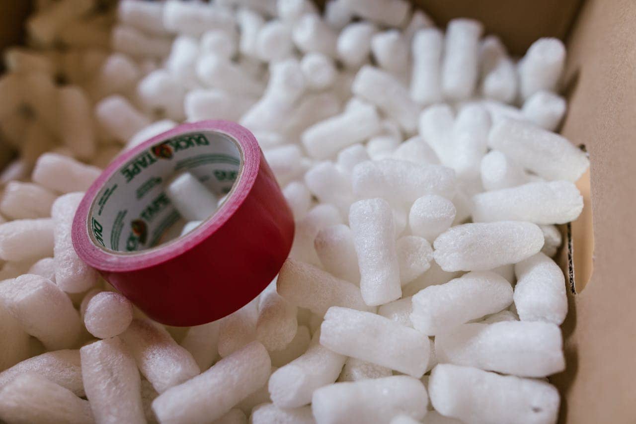 A roll of red duct tape, surrounded by packing peanuts, inside a cardboard box, tape roll partially buried, packing materials scattered