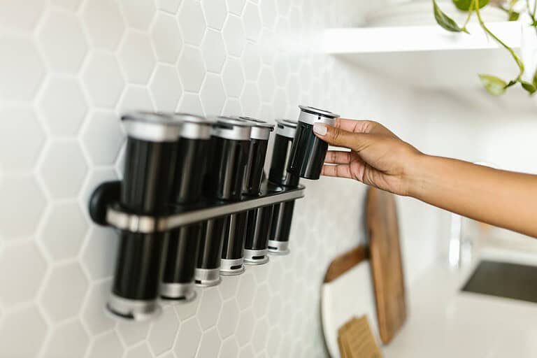 A hand placing a black spice container onto a rack on a hexagonal tiled wall, organized spice rack with multiple containers, modern kitchen setting with a clean, minimalist design, containers with metal lids, kitchen storage solution for spices or herbs