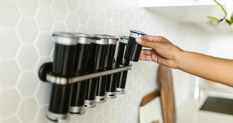 A hand placing a black spice container onto a rack on a hexagonal tiled wall, organized spice rack with multiple containers, modern kitchen setting with a clean, minimalist design, containers with metal lids, kitchen storage solution for spices or herbs
