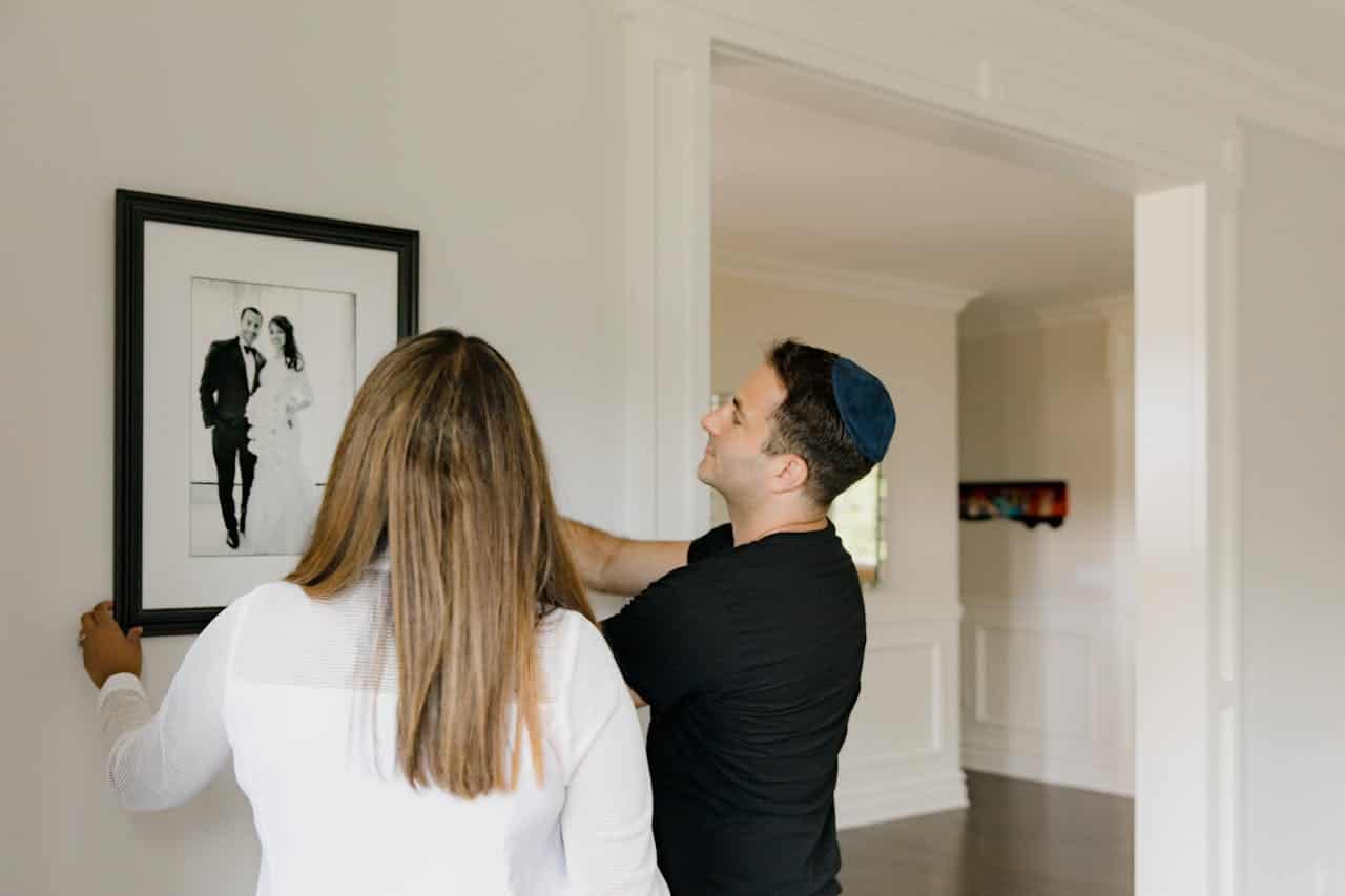 A couple is hanging a framed wedding photo on the wall, the woman is holding the frame while the man adjusts its placement, they are in a bright room with neutral-colored walls, they appear engaged in the task, the man is wearing a kippah, the photo shows the couple in formal wedding attire