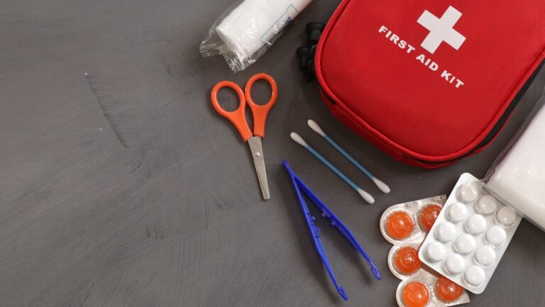 First aid kit with medical supplies including scissors, bandages, cotton swabs, tweezers, and medication on gray surface
