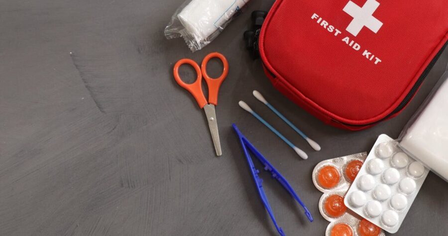 First aid kit with medical supplies including scissors, bandages, cotton swabs, tweezers, and medication on gray surface