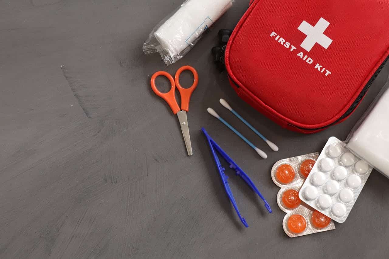First aid kit with medical supplies including scissors, bandages, cotton swabs, tweezers, and medication on gray surface
