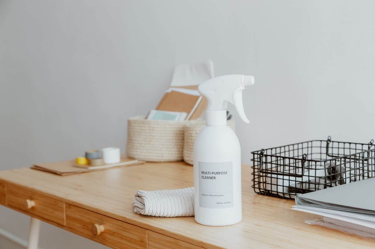A bottle of multi-purpose cleaner placed on a bamboo table, beside a folded cloth, a storage basket, rolls of tape, and paper, items arranged for cleaning or organizing