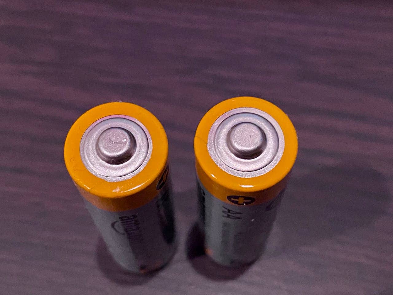 Two AA batteries with yellow tops, standing upright on a dark wooden surface, positive terminals facing upward, metallic contacts reflecting light, slight wear visible on edges