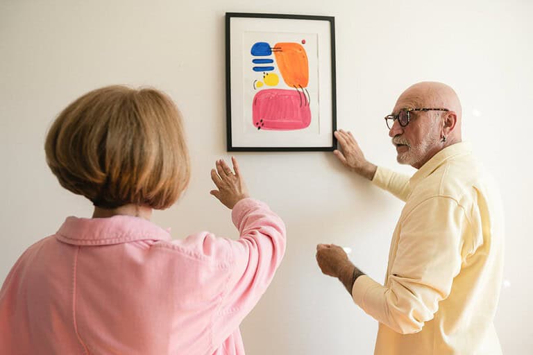 A man and a woman are hanging a framed painting on a wall, the woman is gesturing towards the artwork, while the man is adjusting the frame, they are collaborating in the process of wall decoration