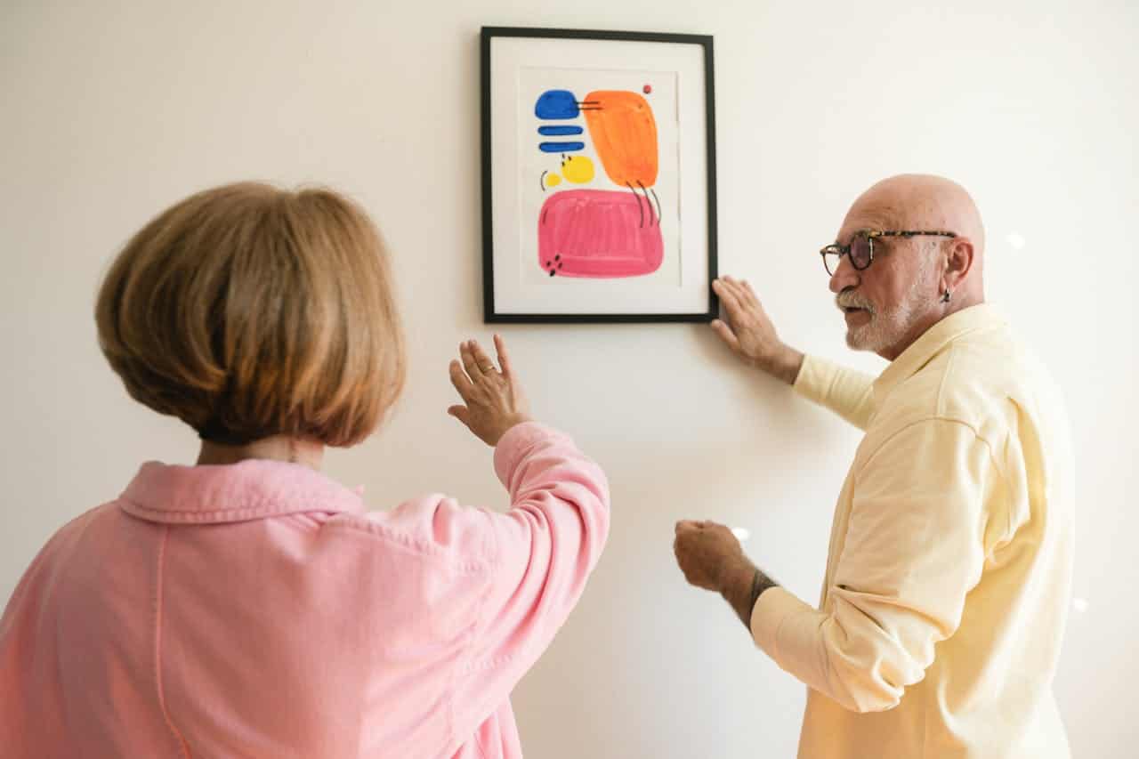 A man and a woman are hanging a framed painting on a wall, the woman is gesturing towards the artwork, while the man is adjusting the frame, they are collaborating in the process of wall decoration