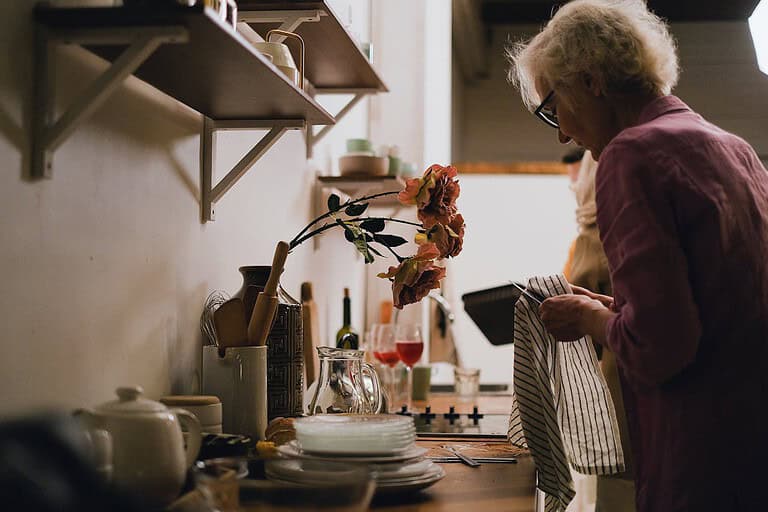 An elderly woman drying dishes in the kitchen, a vase with artificial flowers on the counter, shelves with kitchen utensils and plates
