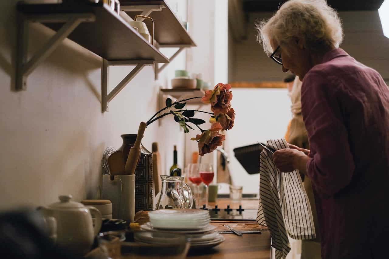 An elderly woman drying dishes in the kitchen, a vase with artificial flowers on the counter, shelves with kitchen utensils and plates
