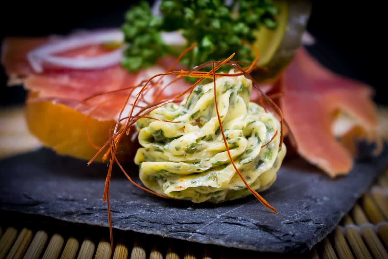 A close-up image of saffron threads placed over a swirl of herb-infused butter, the butter is presented on a black slate plate with thinly sliced prosciutto and a garnish of parsley in the background, the vibrant orange saffron threads contrast with the creamy butter