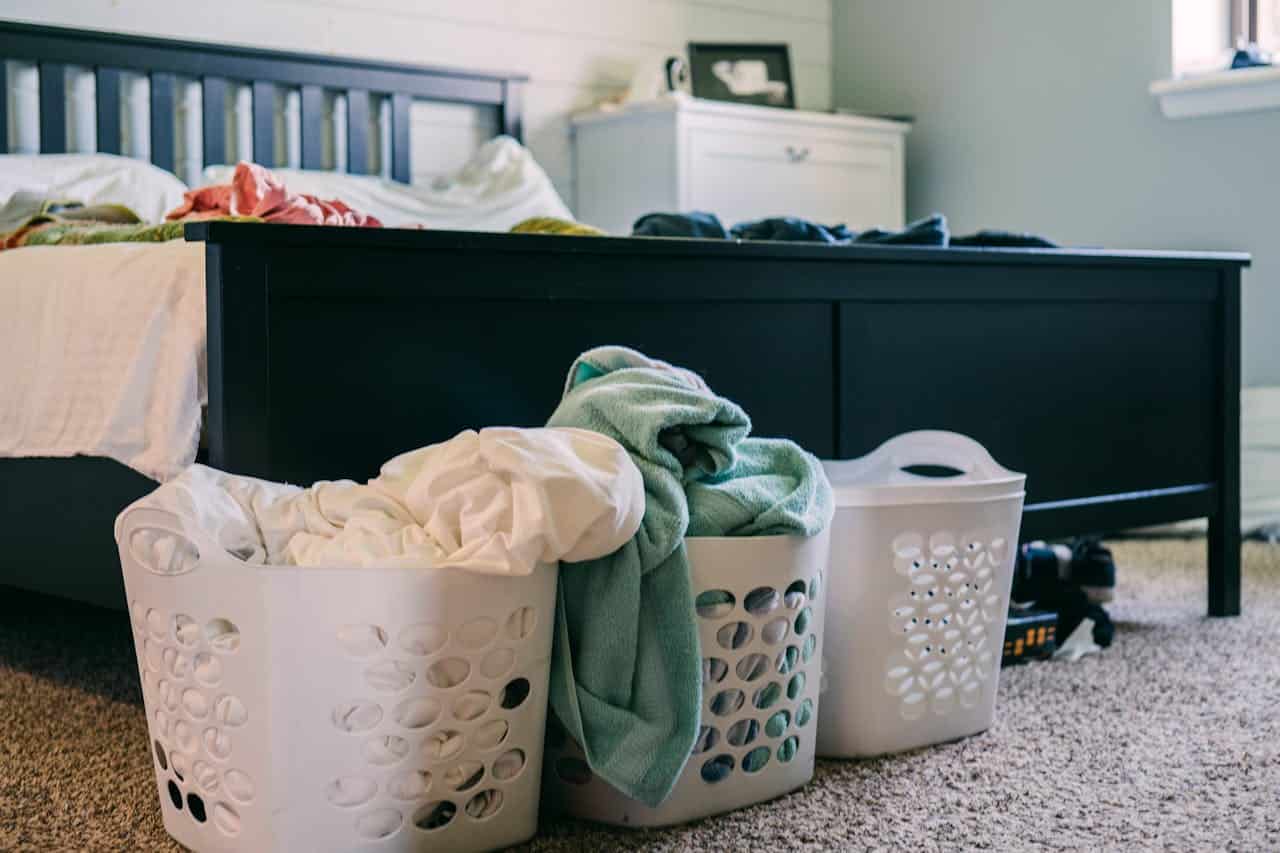 Three white laundry baskets with clothes in bedroom with dark bed frame, pale blue walls and carpeted floor