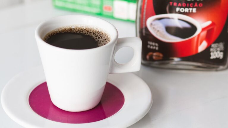A white cup of black coffee sitting on a pink saucer, with frothy coffee visible on top, next to a Nescafé Tradicional Forte coffee jar
