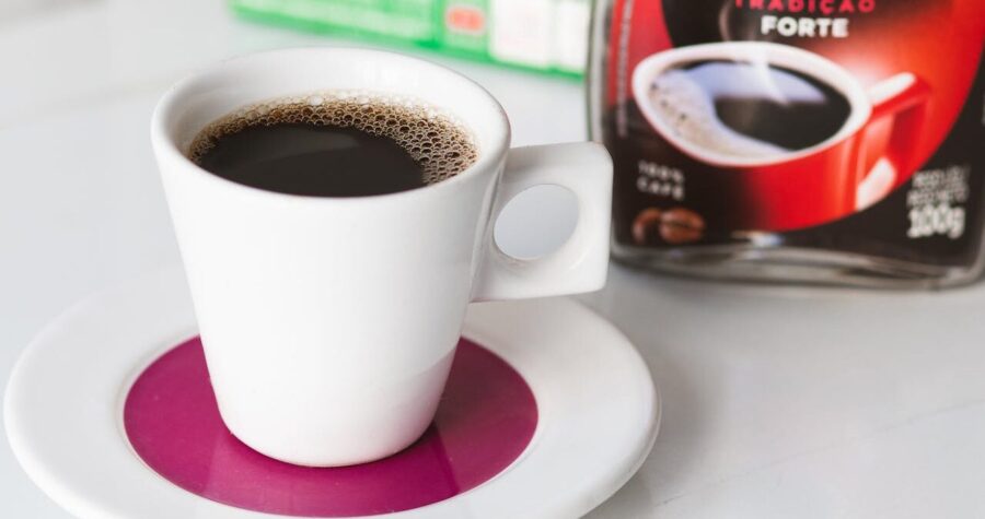 A white cup of black coffee sitting on a pink saucer, with frothy coffee visible on top, next to a Nescafé Tradicional Forte coffee jar