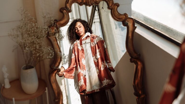 A person standing in front of a decorative mirror, holding a shirt, the reflection shows the individual and the environment, with sunlight streaming through a window, a glimpse of a vase and a wooden table in the background, the mirror's ornate frame adds a vintage touch to the scene