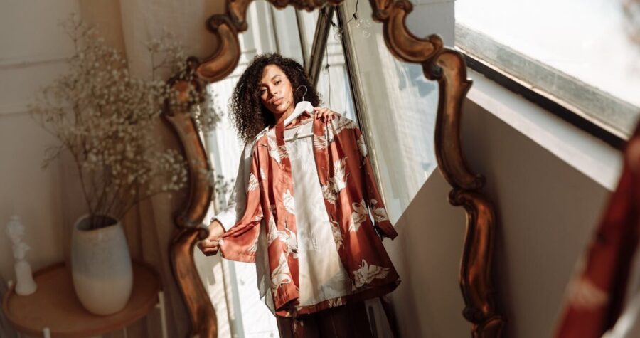 A person standing in front of a decorative mirror, holding a shirt, the reflection shows the individual and the environment, with sunlight streaming through a window, a glimpse of a vase and a wooden table in the background, the mirror's ornate frame adds a vintage touch to the scene